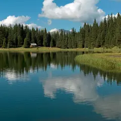 Riflessi di un cielo azzurro e nuvoloso sul lago di Cece, circondato da fitti abeti e una piccola baita.