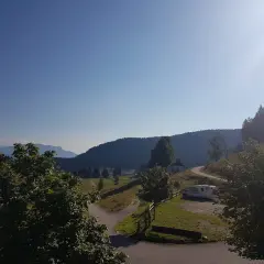 Un camper parcheggiato vicino a un **rifugio barricata** in montagna, con un sentiero che si snoda tra prati verdi e boschi.