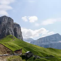 Edifici alpini e un parcheggio affollato ai piedi di imponenti pareti rocciose sul passo Sella, con verdi pendii erbosi.
