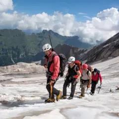 Alpinisti in cordata attraversano un ghiacciaio innevato, con montagne verdi sullo sfondo. Un'avventura al Passo Fedaia.