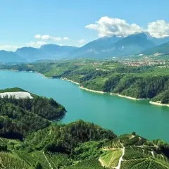 Vista aerea del lago artificiale della diga di Santa Giustina, circondato da rigogliose colline coltivate a vigneti e frutteti, con montagne innevate all'orizzonte.