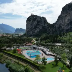Agricamp Altogarda: un villaggio vacanze con piscine e scivoli ai piedi di imponenti montagne, vicino a un fiume e un borgo.