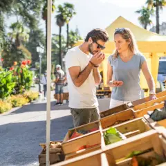 Coppia al markt Riva del Garda, annusando prodotti freschi. Verdure in cassette di legno sotto il sole.