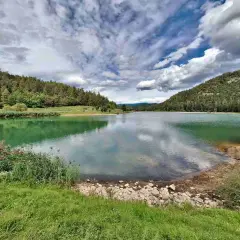 Il lago di Tavon, circondato da boschi verdi, riflette un cielo nuvoloso.