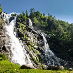 Le Cascate Nardis, un imponente salto d'acqua che scende su rocce scure, circondate da un verde lussureggiante e alberi imponenti sotto un cielo azzurro.