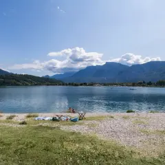 Relaxing day al lido di Caldonazzo, con montagne sullo sfondo e persone che si godono il sole e il lago.