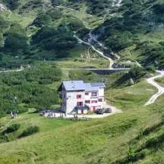 Il rifugio Scalorbi, un edificio in pietra con persiane rosse, si erge in una valle verdeggiante, circondato da sentieri e vegetazione alpina.
