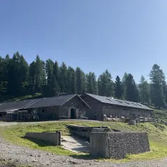 Malga Vallazza, un rifugio alpino in pietra con tetti scuri, circondato da un bosco di abeti sotto un cielo azzurro.