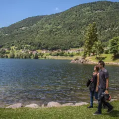 Coppia passeggia sul lungolago del lago delle piazze, con montagne verdi e un villaggio sullo sfondo.