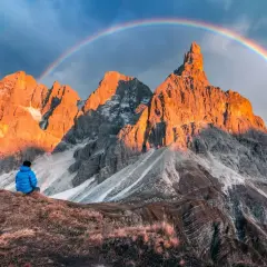 Persona seduta ammira le maestose Pale di San Martino illuminate dal sole al tramonto, con un arcobaleno che le sovrasta.