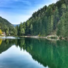 Un **lago smeraldo** incastonato tra montagne boscose, con un ponte pedonale che riflette le sue acque cristalline.