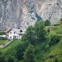 Rifugio Casarota, un'accogliente struttura bianca con persiane blu, incastonata nel verde di un pendio montano, sotto imponenti pareti rocciose.