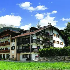 Albergo La Montanara a Predazzo, con le sue balconate in legno e le montagne sullo sfondo, incorniciato da un cielo azzurro e nuvole bianche.