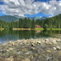 Il lago dei caprioli, circondato da fitti boschi e montagne, riflette un cielo azzurro con nuvole bianche. Una casa in legno si affaccia sull'acqua.