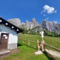 Malga Cavallera: sentiero con cartelli direzionali e rifugio bianco ai piedi delle maestose montagne.