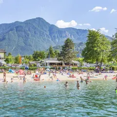 Spiaggia affollata al camping Riviera Caldonazzo, con gente che prende il sole e nuota nel lago, montagne sullo sfondo.