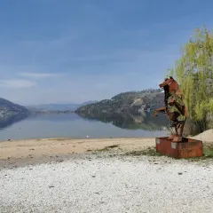 Scultura metallica di un cane sulla riva del lago di Caldonazzo, con montagne riflesse sull'acqua calma.