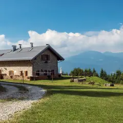 Malga Campo, un rifugio in pietra con tetto metallico, immerso nel verde di un prato alpino, con montagne sullo sfondo.