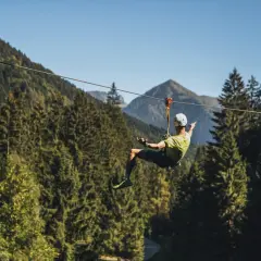 Uomo in volo su una zipline in un parco avventura Trentino, circondato da fitti boschi e montagne.