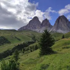 Verdi pendii alpini verso le imponenti cime del Sassolungo, con sentieri e impianti di risalita. Immagina le cascate basaltiche di Canazei in lontananza.