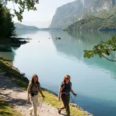 Due escursioniste camminano lungo il sentiero del lago di Molveno in estate, con montagne verdi sullo sfondo.