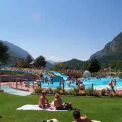Giornata di sole all'acquapark Molveno, con scivoli blu, piscine piene di gente e montagne sullo sfondo.