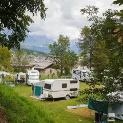 Un tranquillo camping calavise con roulotte e camper immersi nel verde, sullo sfondo un villaggio alpino e montagne nebbiose.