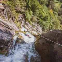 Donna in zipline sopra una cascata nella Val Daone, con rocce e alberi circostanti.