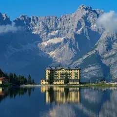 Un hotel giallo si specchia in un lago alpino, con imponenti montagne sullo sfondo: ecco dove si trovano le Dolomiti.