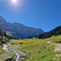 Sentiero sterrato che sale verso un rifugio alpino, con un ruscello che scorre in primo piano e montagne imponenti sullo sfondo, tipico del passo Duron.