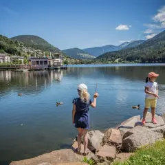 Bambine al lago di Serraia, con montagne verdi e un villaggio sullo sfondo.