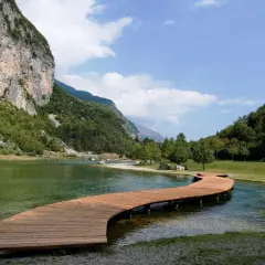 Un pontile in legno serpeggia sul lago di Andalo, circondato da montagne verdi e un cielo azzurro con nuvole bianche.