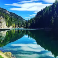 Un lago smeraldo incastonato tra montagne boscose. Una spiaggia erbosa invita al relax, mentre un ponte attraversa le acque calme.