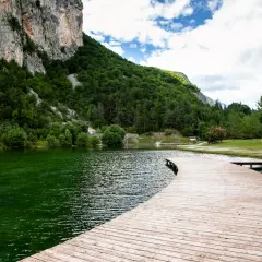 Un pontile in legno curva dolcemente sul lago di Nembia, circondato da montagne verdi e un cielo nuvoloso.