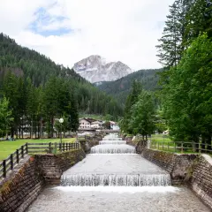 Cascata artificiale nel **Val di Fassa**, con montagne boscose e un villaggio sullo sfondo.
