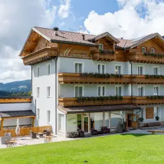 L'albergo Miramonti, con le sue balconate fiorite e il tetto in legno, si staglia contro un cielo azzurro con nuvole bianche, immerso in un paesaggio montano verdeggiante.