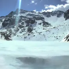 Il lago delle stellune, ghiacciato e circondato da montagne innevate, riflette il cielo azzurro e le nuvole.