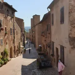 Una persona con mascherina passeggia per una stretta via acciottolata di **Certaldo Alto**, circondata da antichi edifici in mattoni con persiane in legno.