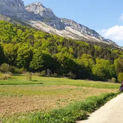 Ciclisti su un sentiero sterrato, con campi coltivati e un bosco rigoglioso ai piedi di una montagna imponente. Un passo bordala da godersi in bicicletta.