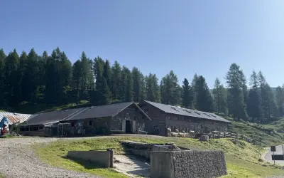 Malga Vallazza, un rifugio alpino in pietra con tetti scuri, circondato da un bosco di abeti sotto un cielo azzurro.