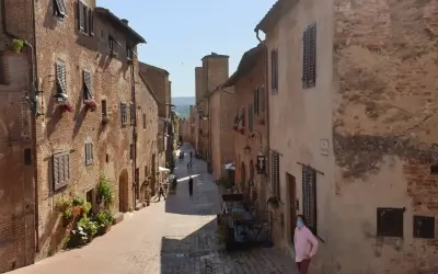 Una persona con mascherina passeggia per una stretta via acciottolata di **Certaldo Alto**, circondata da antichi edifici in mattoni con persiane in legno.