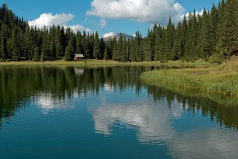 Riflessi di un cielo azzurro e nuvoloso sul lago di Cece, circondato da fitti abeti e una piccola baita.