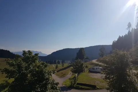 Un camper parcheggiato vicino a un **rifugio barricata** in montagna, con un sentiero che si snoda tra prati verdi e boschi.