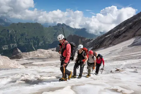 Alpinisti in cordata attraversano un ghiacciaio innevato, con montagne verdi sullo sfondo. Un'avventura al Passo Fedaia.