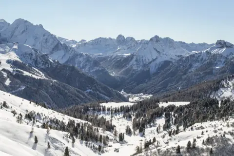 Panorami innevati di Canazei in inverno, con montagne maestose, boschi di conifere e un villaggio nascosto nella valle.