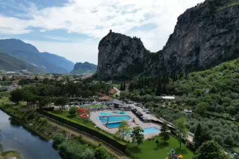 Agricamp Altogarda: un villaggio vacanze con piscine e scivoli ai piedi di imponenti montagne, vicino a un fiume e un borgo.