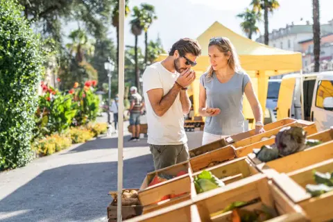 Coppia al markt Riva del Garda, annusando prodotti freschi. Verdure in cassette di legno sotto il sole.