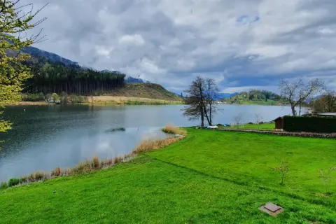 Un tranquillo lago di Pinè con verdi prati, alberi spogli e nuvole minacciose nel cielo.