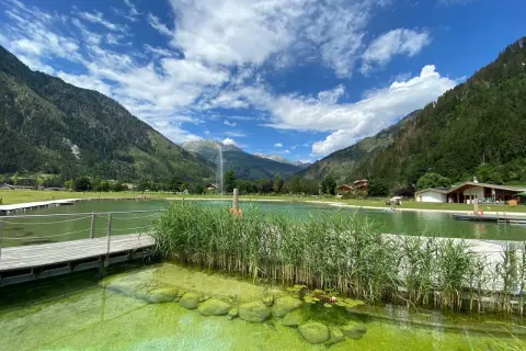 Un rinfrescante biolago a Predazzo, circondato da montagne verdi e un cielo azzurro con nuvole bianche. Pontili in legno e canneti creano un'atmosfera serena.