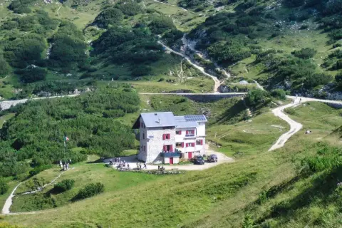 Il rifugio Scalorbi, un edificio in pietra con persiane rosse, si erge in una valle verdeggiante, circondato da sentieri e vegetazione alpina.